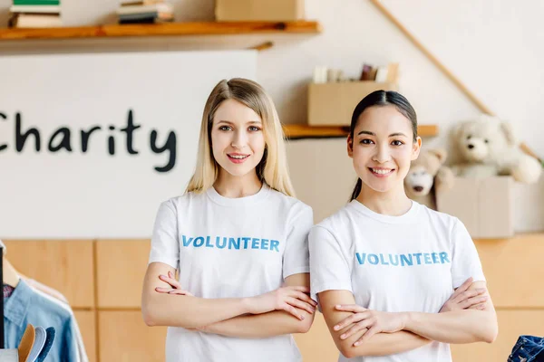 Dos hermosas chicas voluntarias multiculturales con los brazos cruzados sonriendo y mirando a la cámara - foto de stock