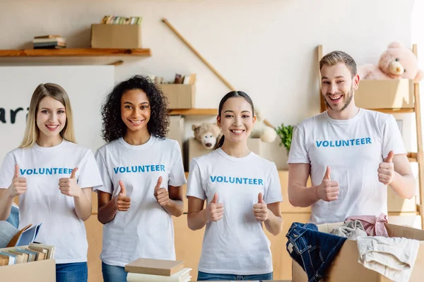 Grupo de quatro jovens voluntários sorridentes em camisetas brancas com inscrições voluntárias mostrando polegares para cima — Fotografia de Stock