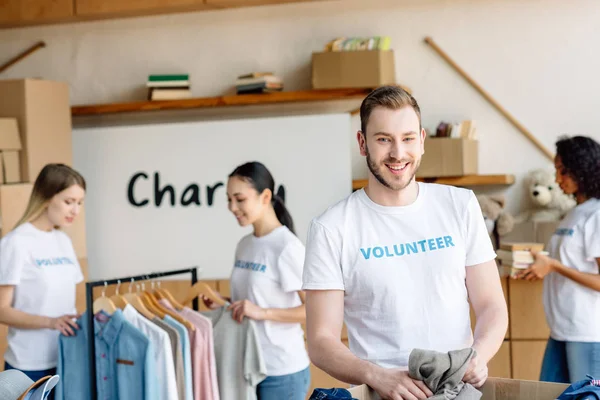 Enfoque selectivo de guapo voluntario desembalaje caja de cartón con ropa cerca de las niñas multiculturales que trabajan en el centro de caridad - foto de stock