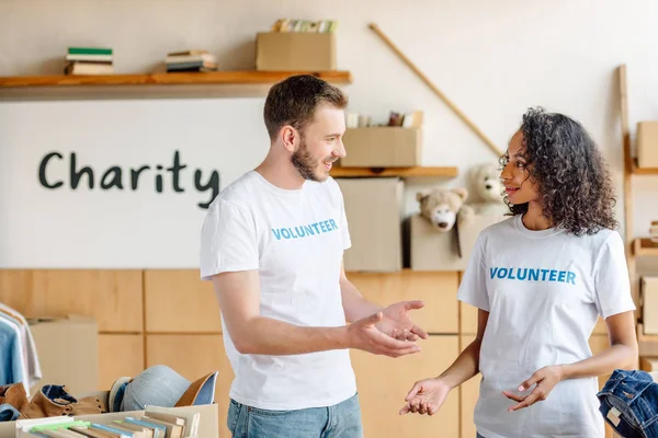 Two multicultural volunteers talking while standing near carton boxes with books and clothes — Stock Photo