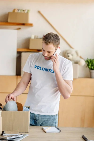 Guapo voluntario hablando en el teléfono inteligente mientras está de pie cerca de caja de cartón con ropa - foto de stock