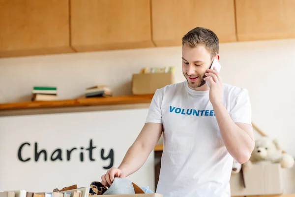 Apuesto joven voluntario hablando en el teléfono inteligente, mientras que de pie cerca de la caja de cartón con ropa - foto de stock