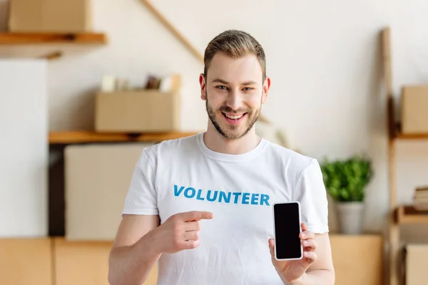 Sonriente joven voluntario apuntando con el dedo al teléfono inteligente con pantalla en blanco - foto de stock