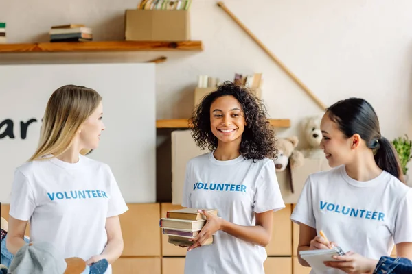 Pretty, multicultural girls in white t-shirts with volunteer inscriptions smiling and looking at each other — Stock Photo