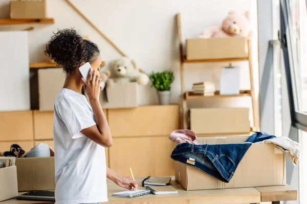 Seitenansicht eines jungen afrikanisch-amerikanischen Freiwilligen, der auf dem Smartphone spricht und in ein Notizbuch schreibt — Stockfoto