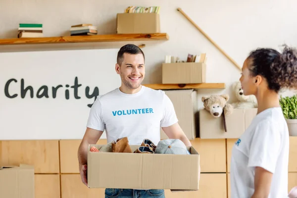 Selective focus of cheerful volunteer holding carton box with clothes near african american girl — Stock Photo