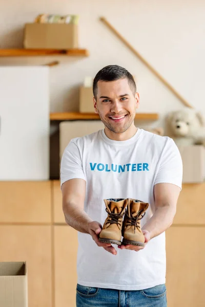 Bonito, sorrindo voluntário segurando sapatos infantis e olhando para a câmera — Fotografia de Stock