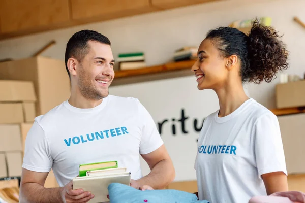 Zwei fröhliche multikulturelle Freiwillige unterhalten sich, während sie Bücher und Kleidung in der Hand halten — Stockfoto