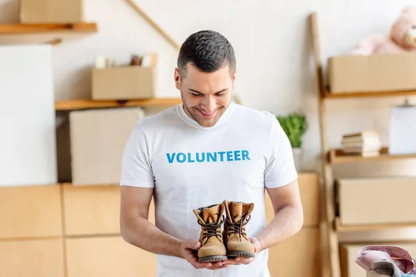 Handsome, young man in white t-shirt with volunteer inscription holding kids shoes — Stock Photo