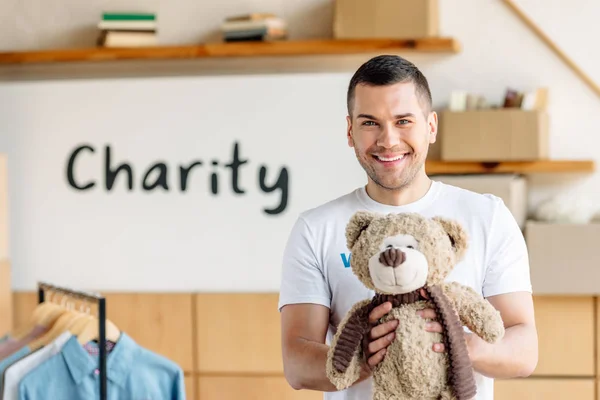 Guapo, alegre voluntario sosteniendo osito de peluche y mirando a la cámara - foto de stock