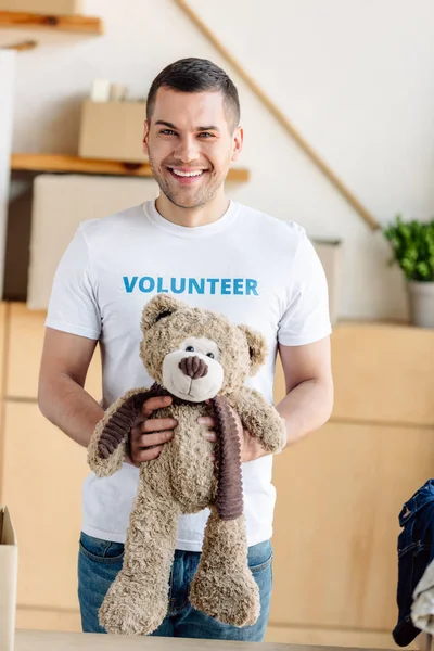 Bien parecido, voluntario sonriente sosteniendo oso de peluche y mirando a la cámara - foto de stock