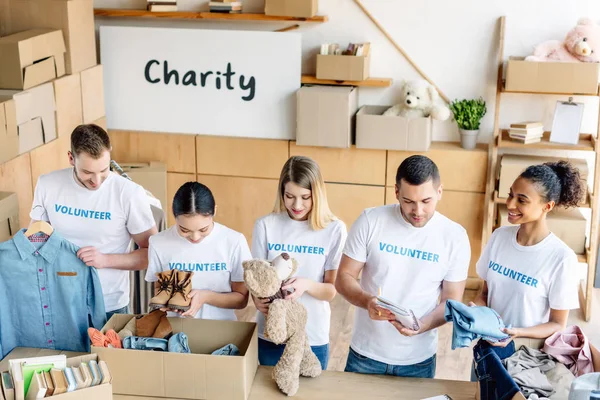 Grupo de jóvenes voluntarios multiculturales desempacando cajas de cartón con ropa, libros y juguetes - foto de stock