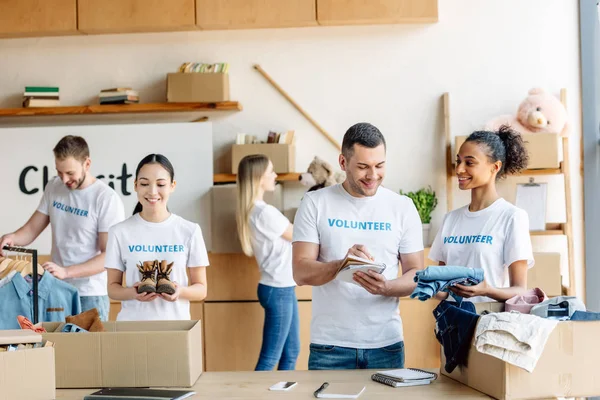 Group of young multicultural volunteers in white t-shirts with volunteer inscriptions working in charity center — Stock Photo