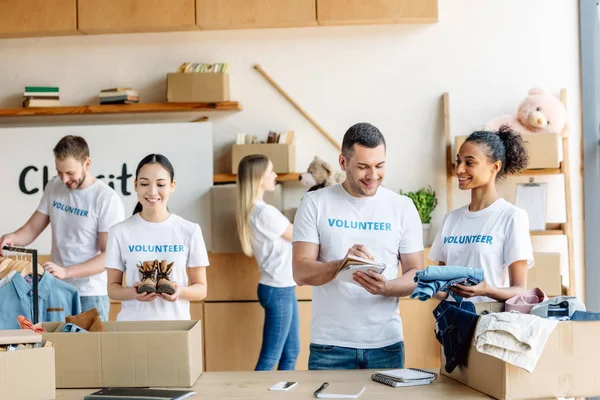 Groupe de jeunes bénévoles multiculturels en t-shirts blancs avec inscriptions bénévoles travaillant dans un centre caritatif — Photo de stock