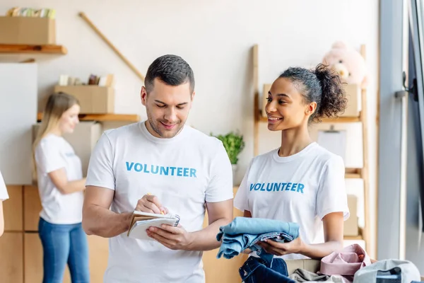 Enfoque selectivo de voluntarios multiculturales sonrientes escribiendo en cuaderno y desempacando caja con ropa - foto de stock