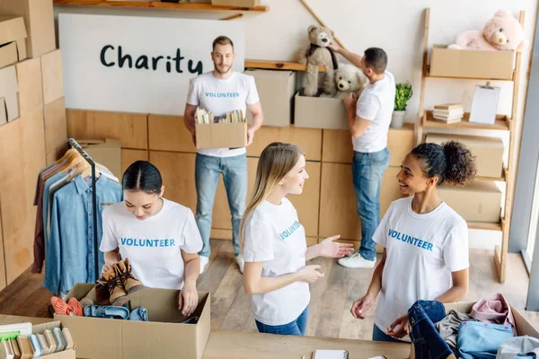 Grupo de jóvenes voluntarios multiculturales en camisetas blancas con inscripciones de voluntarios que trabajan en el centro de caridad - foto de stock