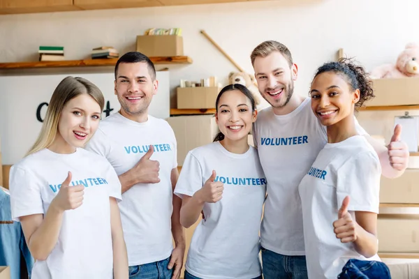 Cinco jóvenes voluntarios multiculturales mostrando los pulgares hacia arriba, sonriendo y mirando a la cámara - foto de stock