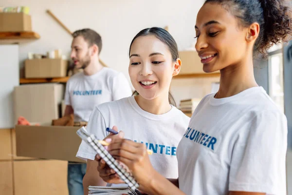 Enfoque selectivo de sonreír a voluntarios multiculturales hablando y escribiendo en un cuaderno - foto de stock