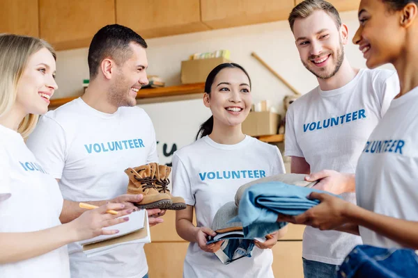 Cinque giovani volontari multiculturali che parlano mentre tengono vestiti in un centro di beneficenza — Foto stock