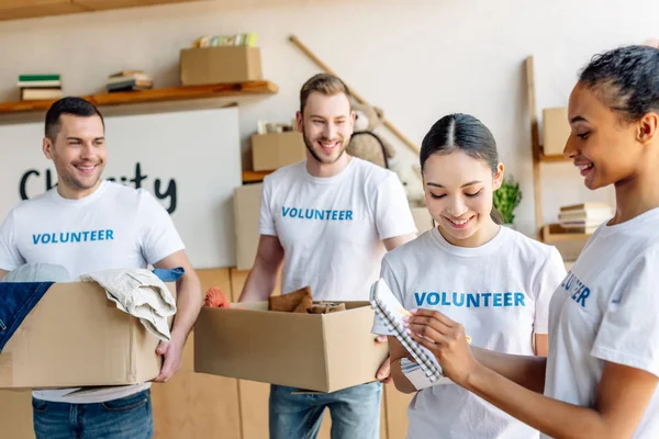 Quattro giovani volontari multiculturali in t-shirt bianche con iscrizioni volontarie che lavorano nel centro di beneficenza — Foto stock