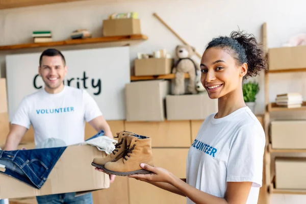 Foco seletivo de muito africano americano voluntário segurando crianças sapatos perto de homem bonito com caixa de papelão — Fotografia de Stock