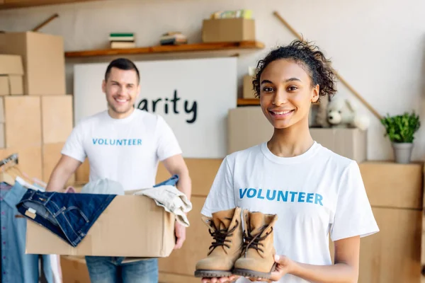 Foco seletivo de menina americana africana bonita segurando crianças sapatos perto de voluntário bonito com caixa de papelão — Fotografia de Stock