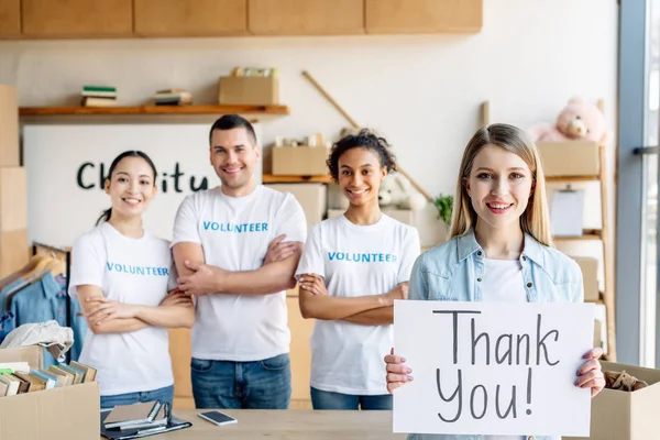 Selektiver Fokus eines hübschen Mädchens mit einem Plakat mit Danksagungstext, während es in der Nähe von multikulturellen Freiwilligen in einem Charity-Zentrum steht — Stockfoto