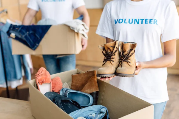 Vista recortada de voluntarios multiculturales desempacando cajas de cartón con ropa y calzado - foto de stock