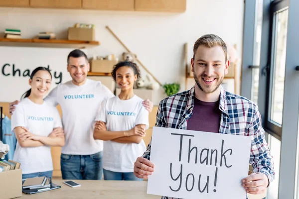 Selective focus of handsome man holding placard with thank you text while standing near multicultural volunteers in charity center — Stock Photo