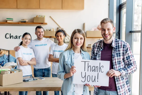 Enfoque selectivo de sonriente hombre y mujer con tarjeta de agradecimiento de texto, mientras que de pie cerca de voluntarios multiculturales alegres en el centro de caridad - foto de stock