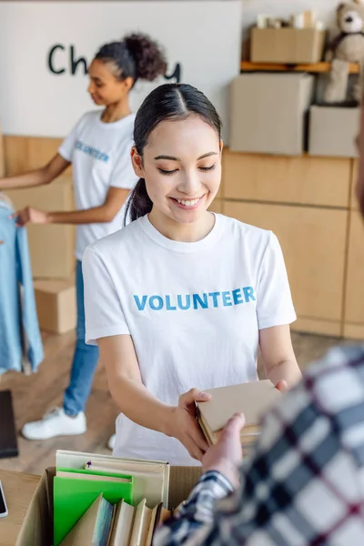 Foco seletivo de sorrir asiático voluntário dando livros para o homem enquanto Africano americano menina de pé perto rack com camisas — Fotografia de Stock