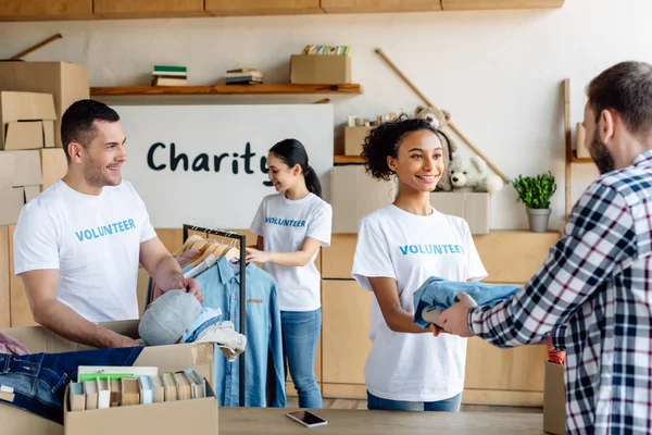 Selective focus of pretty african american girl giving clothes to man while multicultural volunteers unpacking clothes — Stock Photo