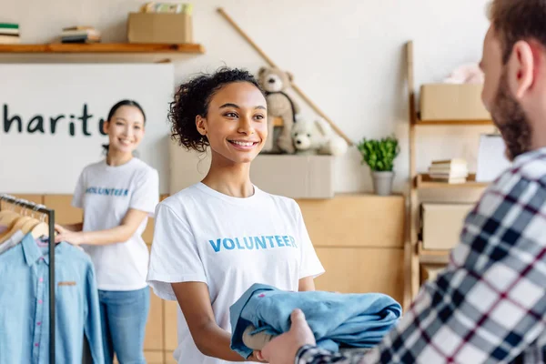 Selective focus of beautiful african american volunteer giving clothes to man while asian girl standing near rack with shirts — Stock Photo