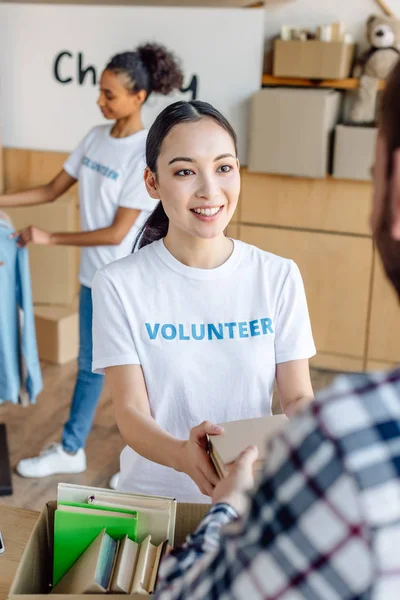 Selective focus of beautiful asian girl giving books to man while african american volunteer working in charity center — Stock Photo