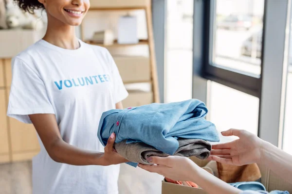 Vista cortada de Africano americano voluntário dando roupas para a mulher no centro de caridade — Fotografia de Stock