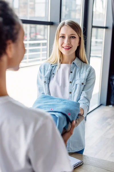 Selective focus of pretty blonde woman taking clothes from african american volunteer — Stock Photo