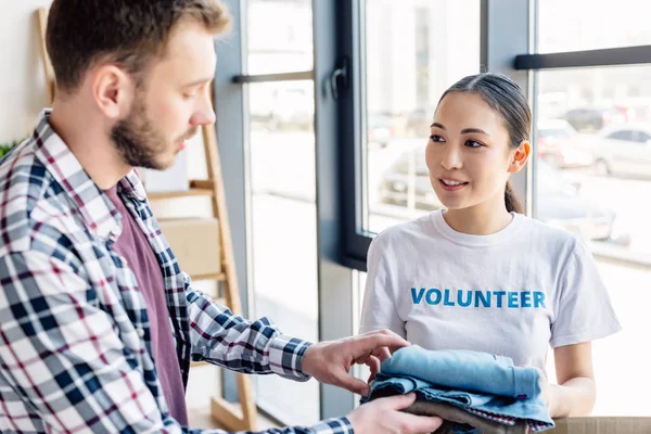 Selective focus of pretty asian volunteer giving clothes to man in charity center — Stock Photo