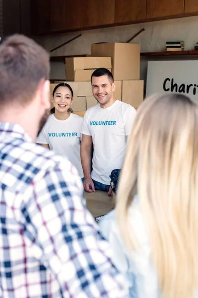 Избирательный фокус улыбающихся мультикультурных волонтеров, встречающих людей в благотворительном центре — стоковое фото