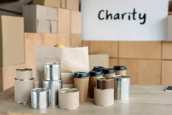 Canned food, disposable cups and paper bag on wooden table in charity center — Stock Photo