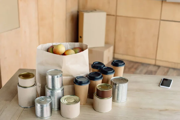 Canned food, disposable cups and paper bag with apples on wooden table in charity center — Stock Photo