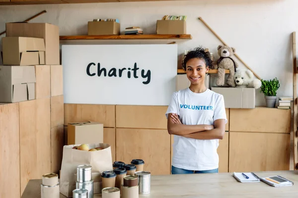 Focus selettivo di sorridente volontario afro-americano in piedi vicino al tavolo con cibo in scatola e bicchieri di carta — Foto stock