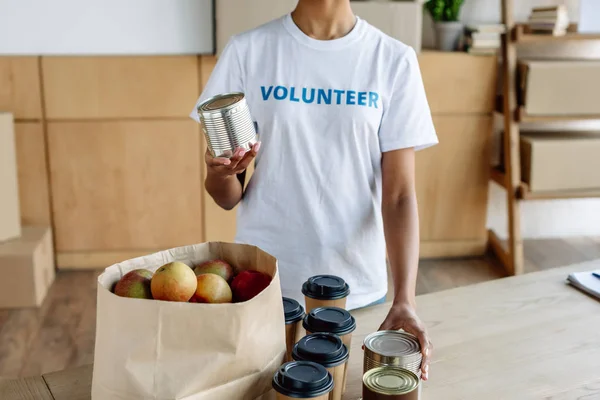 Cropped view of african american volunteer holding tin while standing near table with canned food and paper bag with apples — Stock Photo