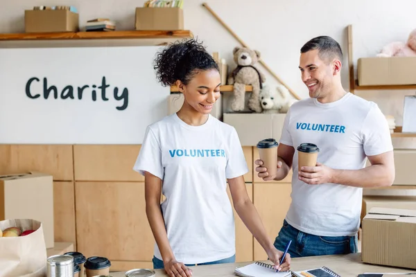 Handsome smiling volunteer holding paper cups while standing near african american girl writing in notebook — Stock Photo