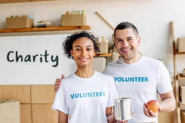 A jóvenes voluntarios multiculturales sonrientes sosteniendo hojalata y manzana y mirando a la cámara - foto de stock