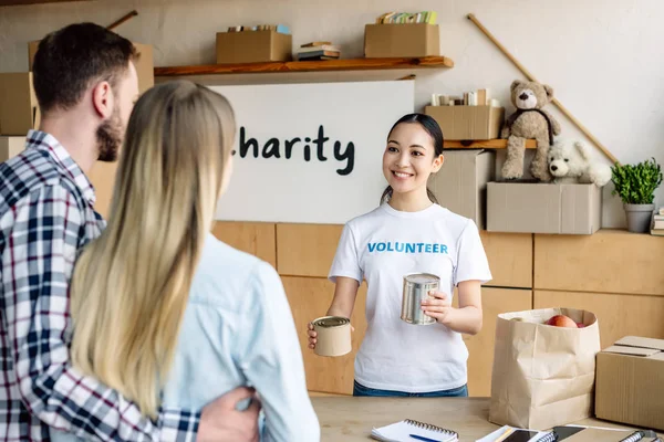 Beautiful asian volunteer girl giving canned food to man and woman in charity center — Stock Photo