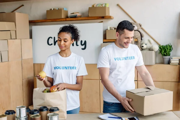 Pretty african american girl putting apples into paper bag while handsome volunteer holding carton box — Stock Photo