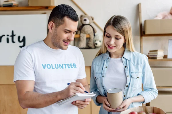 Bonito jovem voluntário escrever no notebook enquanto pé perto bonita mulher loira — Fotografia de Stock