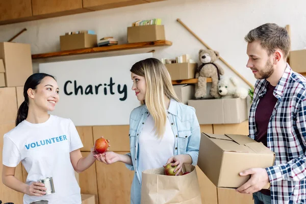 Smiling asian volunteer giving canned food and apples to man and woman in charity center — Stock Photo