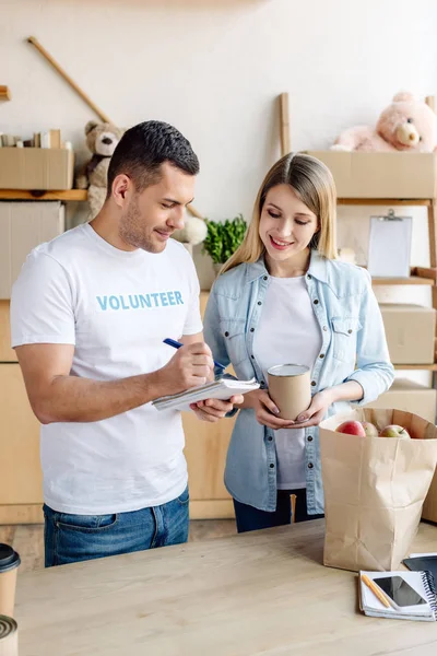 Handsome volunteer writing in notebook while standing near pretty woman holding tin — Stock Photo