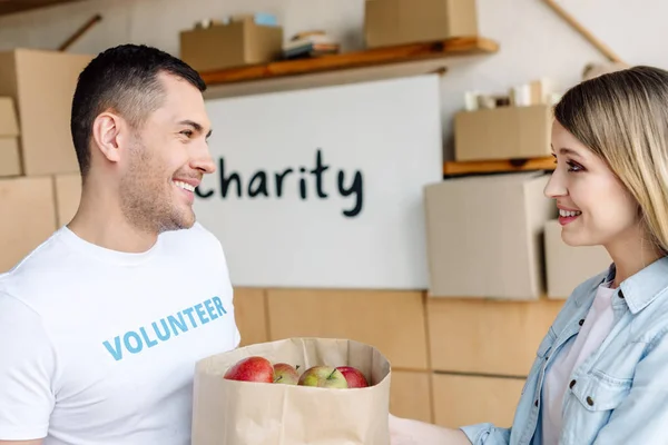 Guapo, voluntario sonriente dando bolsa de papel con manzanas a mujer atractiva - foto de stock